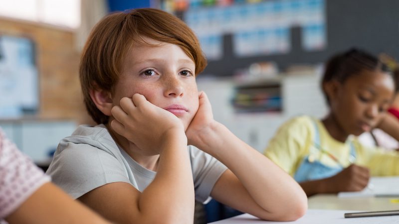 Benefits of Boredom: Tired school boy with hand on face sitting at desk in classroom. Bored schoolchild sitting at desk with classmates in classroom. Frustrated and thoughtful young child sitting and looking up.